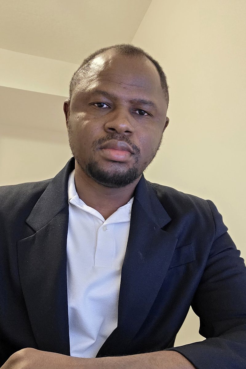 Sadisu Hassan in a navy blazer and white shirt, seated at a desk with a keyboard